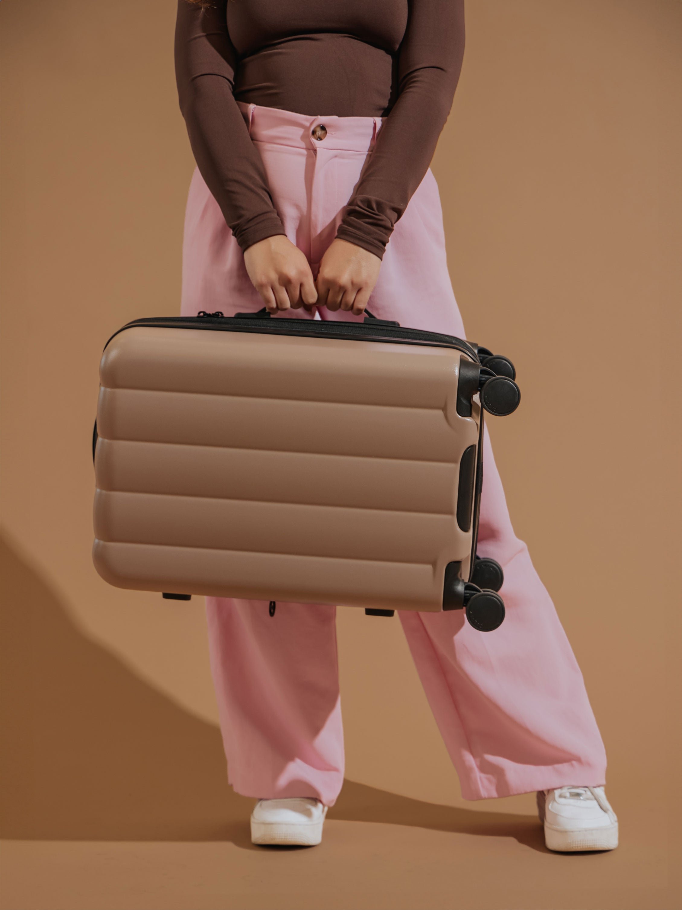 Model holding CALPAK Evry Carry-On Luggage by the side handle in brown
