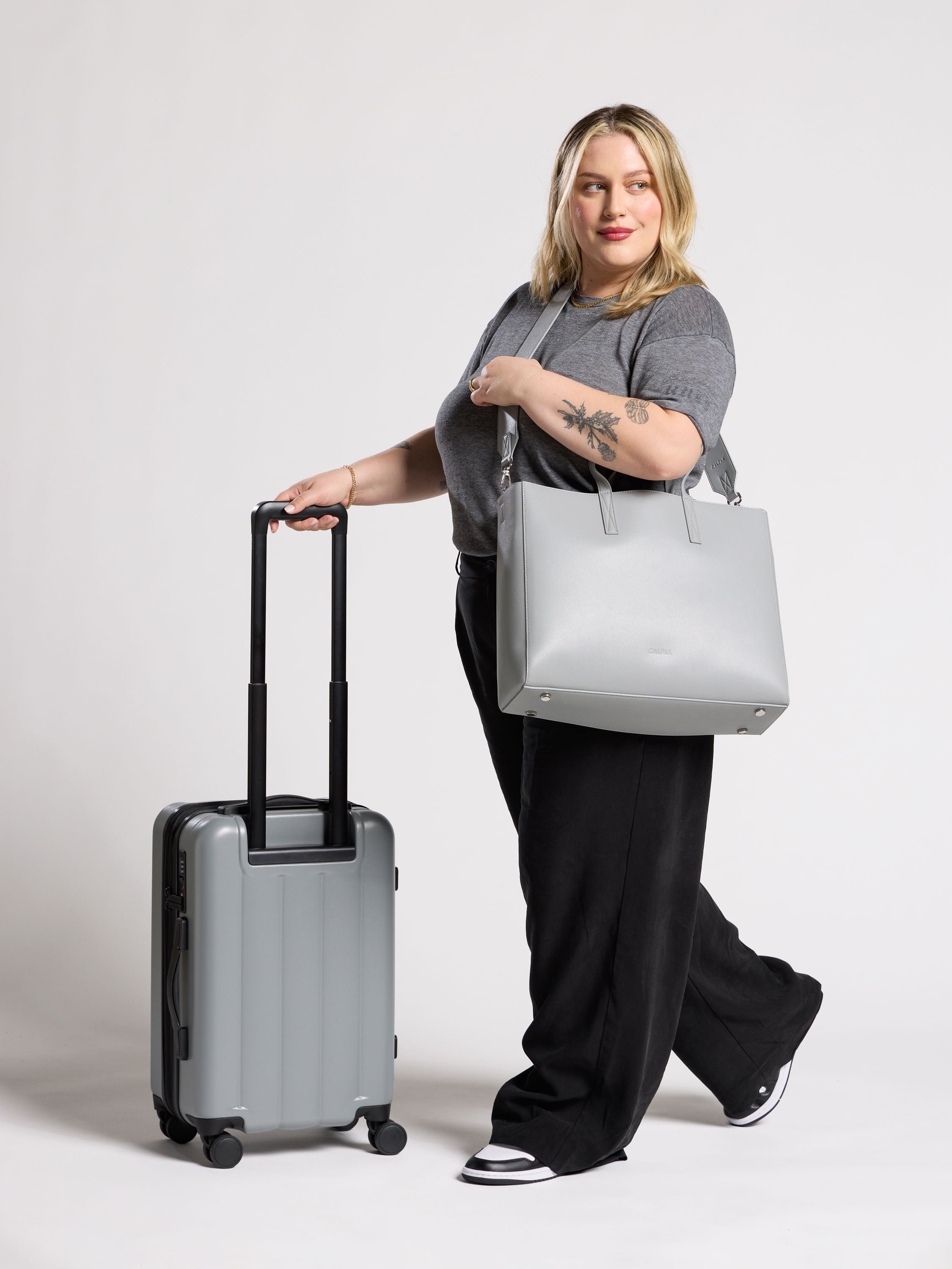 Model wearing CALPAK Haven Laptop Tote over shoulder while rolling Evry Carry-On Luggage in smoke