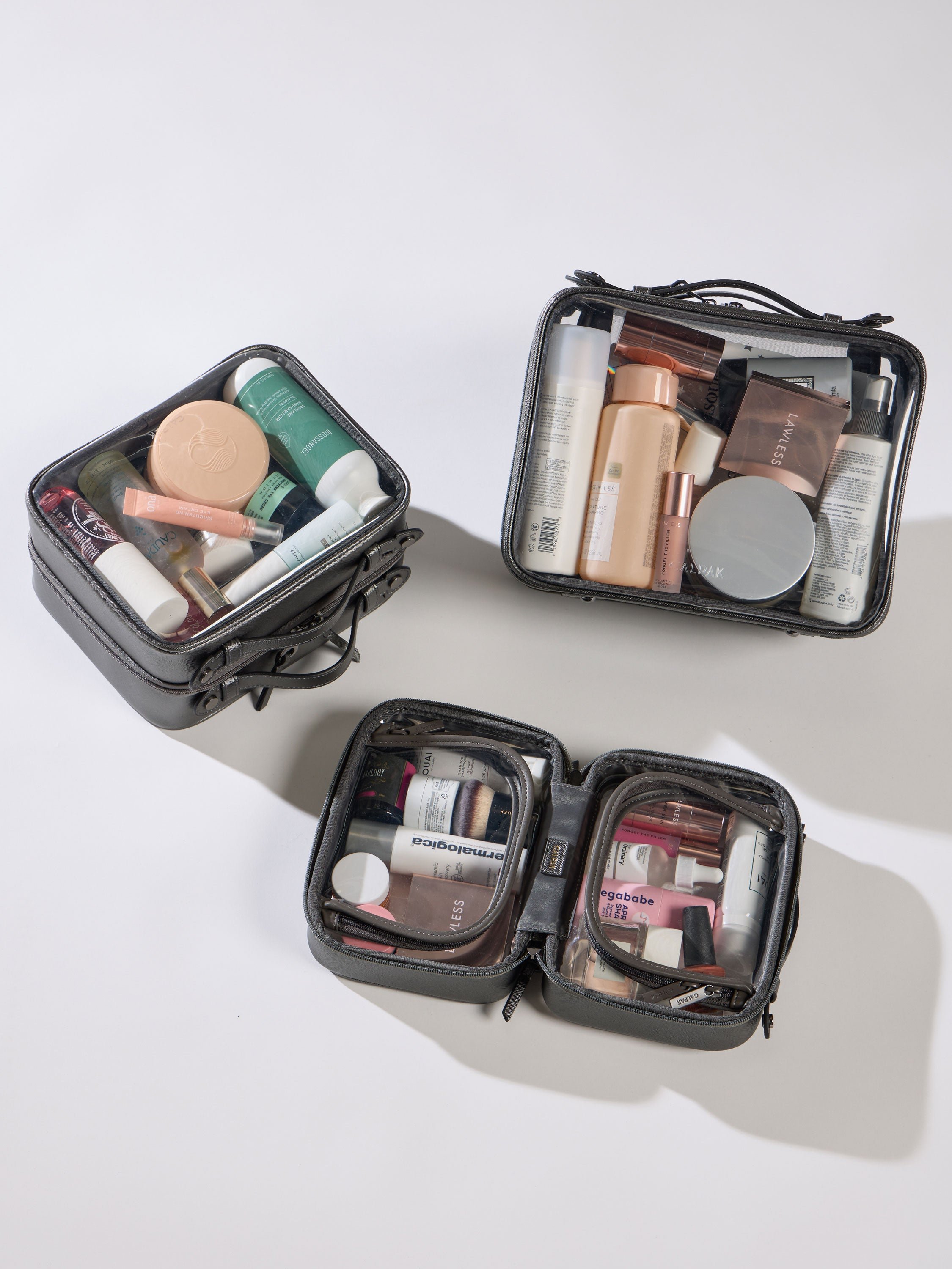 CALPAK Large Clear Cosmetic Case next to Medium Clear Cosmetic Case and Small Clear Cosmetic Case in metallic dark gray