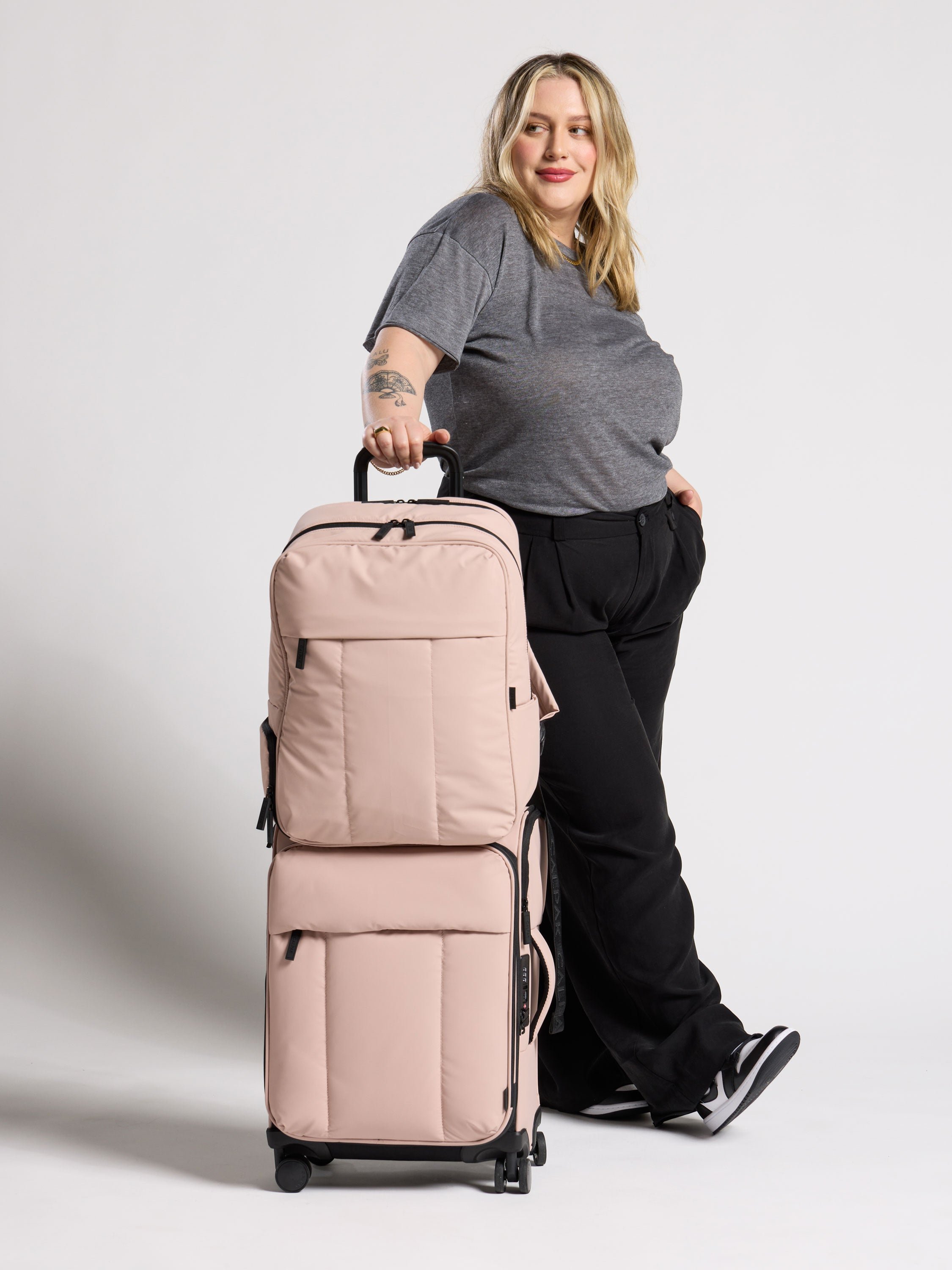 Model standing besides CALPAK 17 inch Laptop Backpack secured to Luka Soft-Sided Carry-On Luggage by trolley pass through in rose quartz
