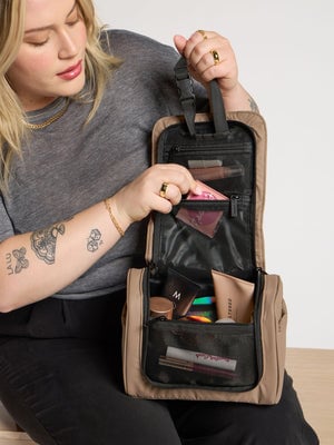 Model placing makeup inside mesh interior pocket of chocolate luka hanging toiletry bag; TLH2301-CHOCOLATE