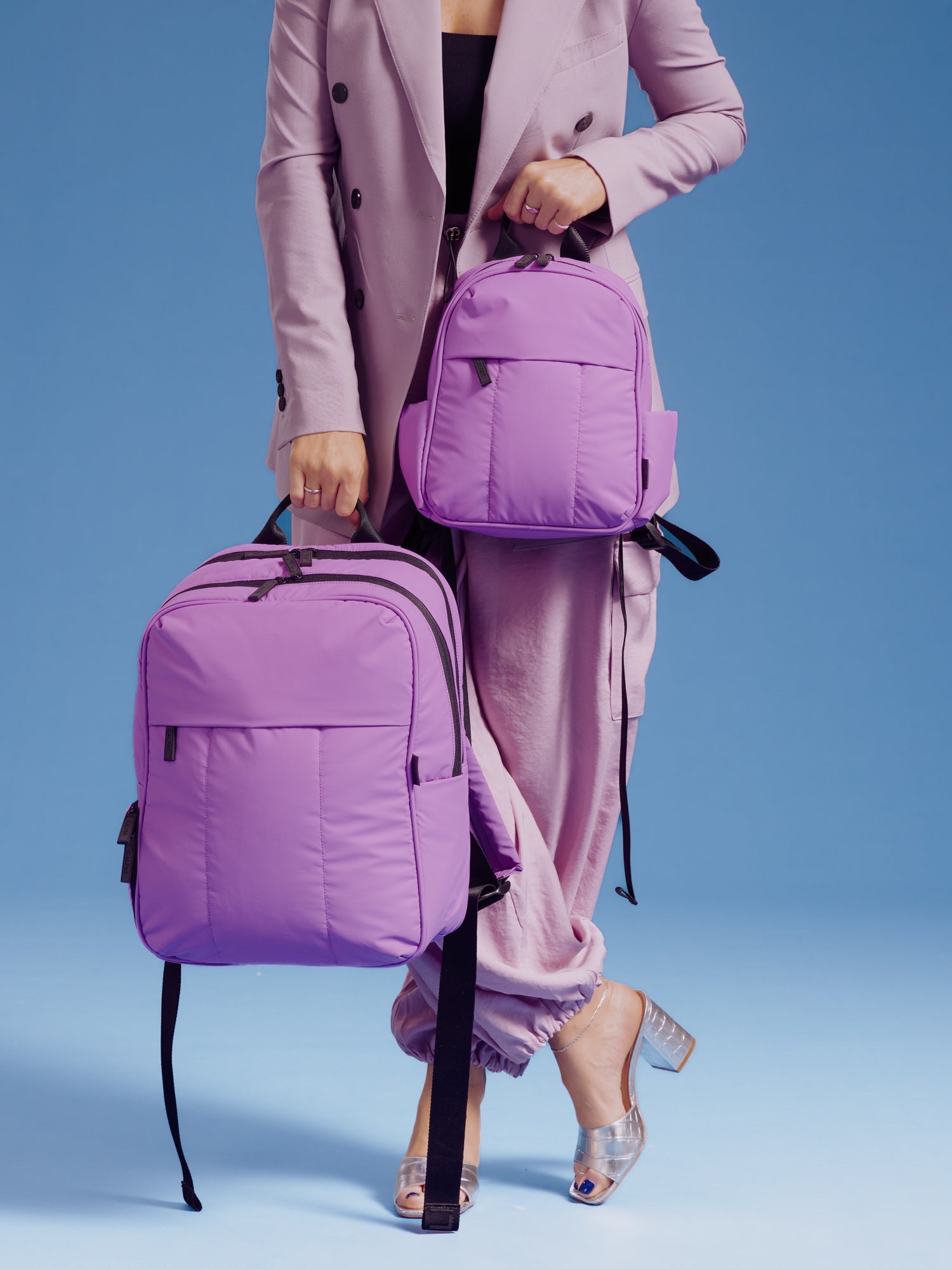 Model holding the CALPAK Luka Mini Backpack and CALPAK 15 inch Laptop Backpack in lavender lilac for size comparison