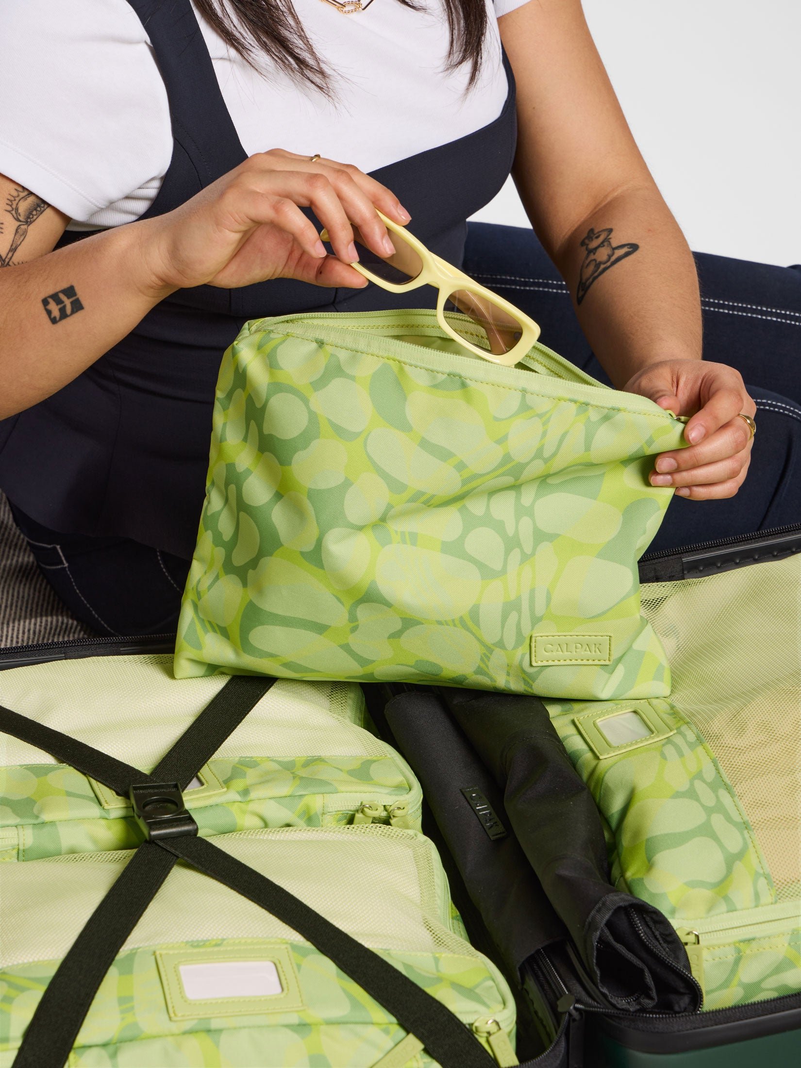Model placing sunglasses in zippered pouch part of CALPAK Packing Cubes 5-Piece Set in green with viper print