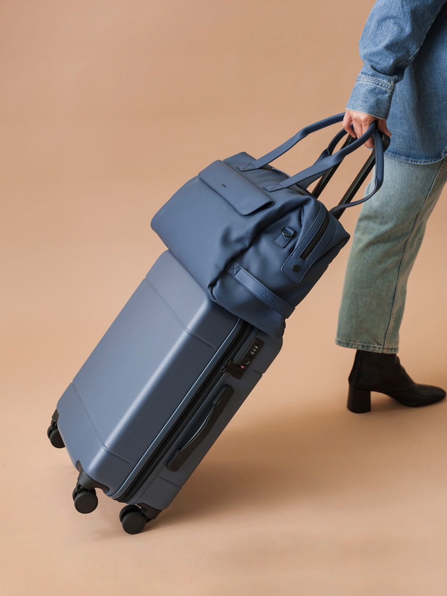 male model holding CALPAK Hue hard shell carry-on suitcase with spinner wheels in blue atlantic color