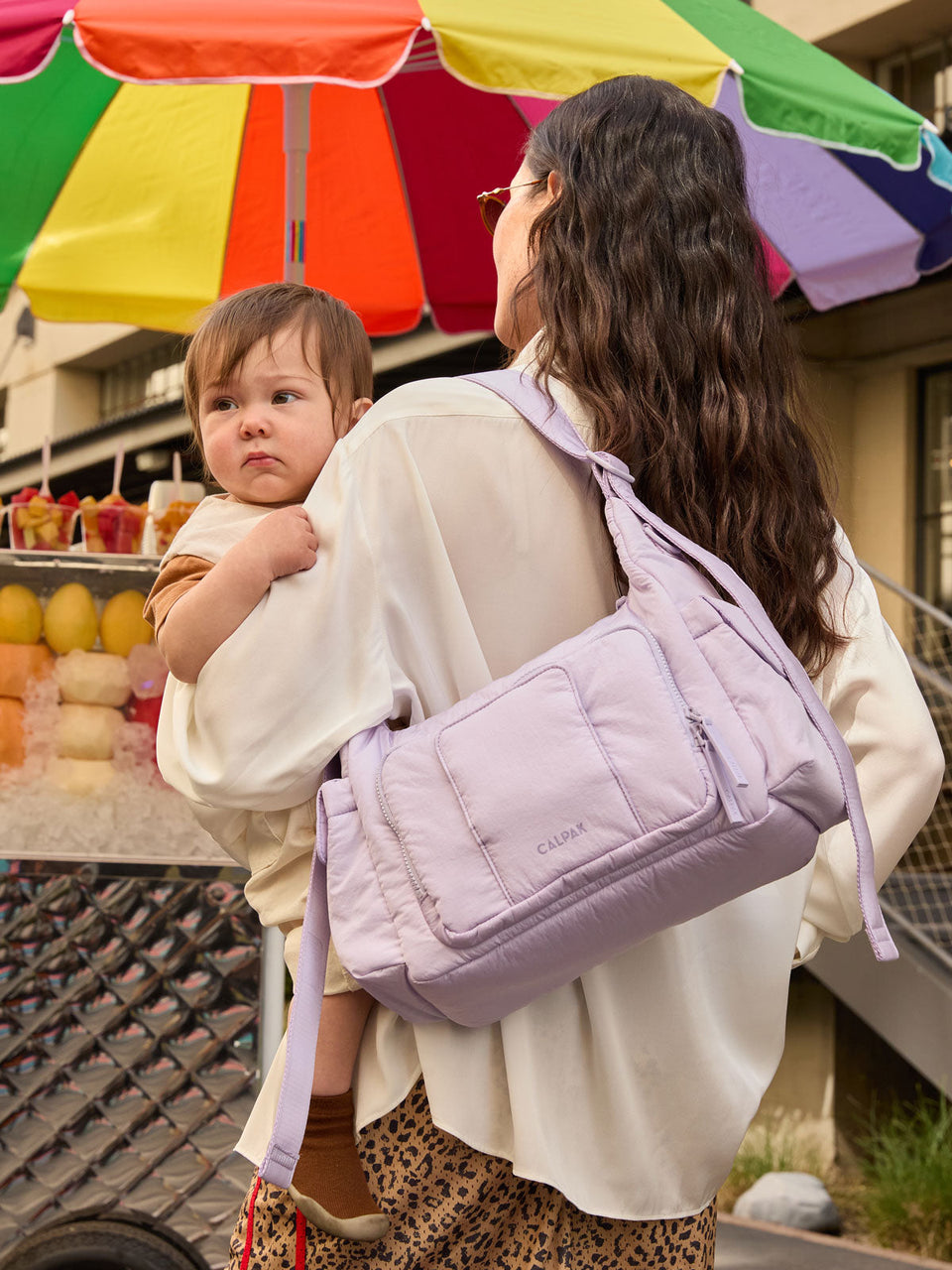 Woman at a bodega with baby in her arms wearing the CALPAK Crossbody Diaper Bag in purple Orchid
