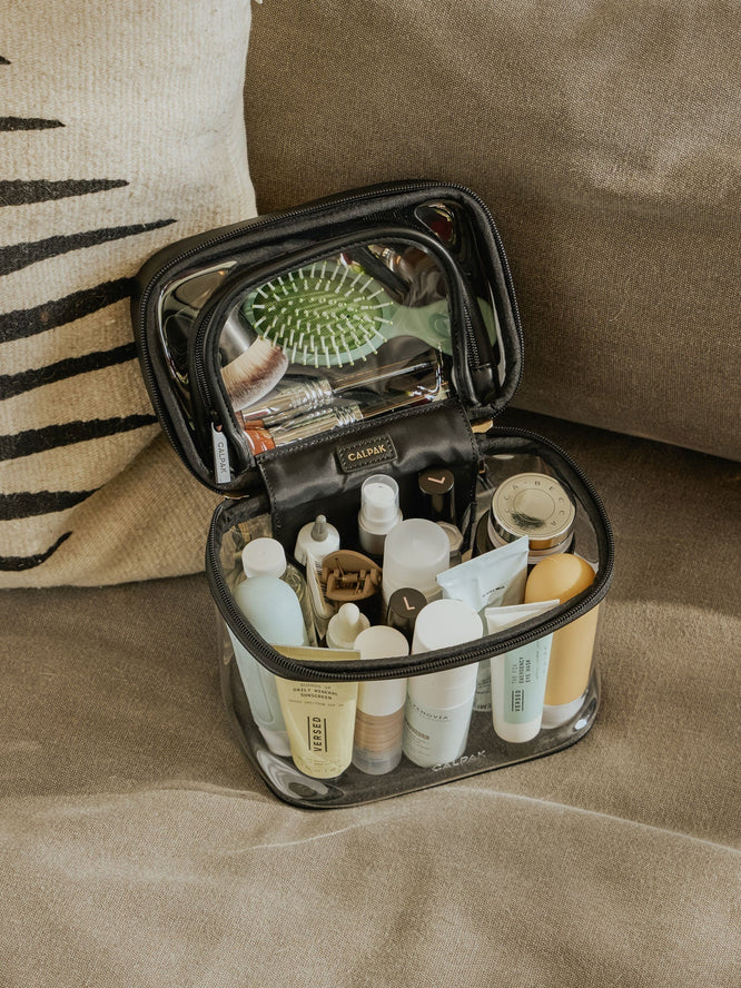 CALPAK Clear train case in black packed with skincare and makeup on a beige couch
