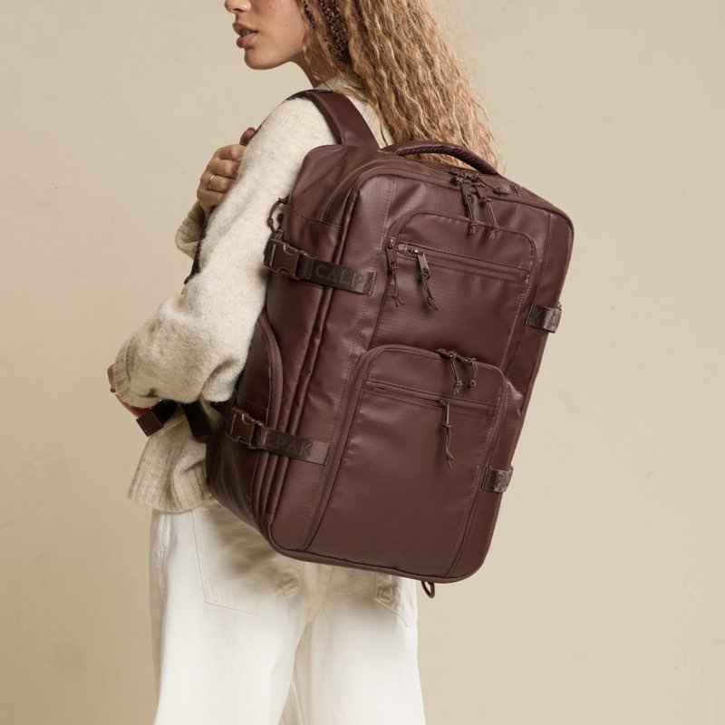 Woman in a beige sweater wearing the CALPAK Terra 26L Laptop Duffel Backpack in brown Walnut against a tan background