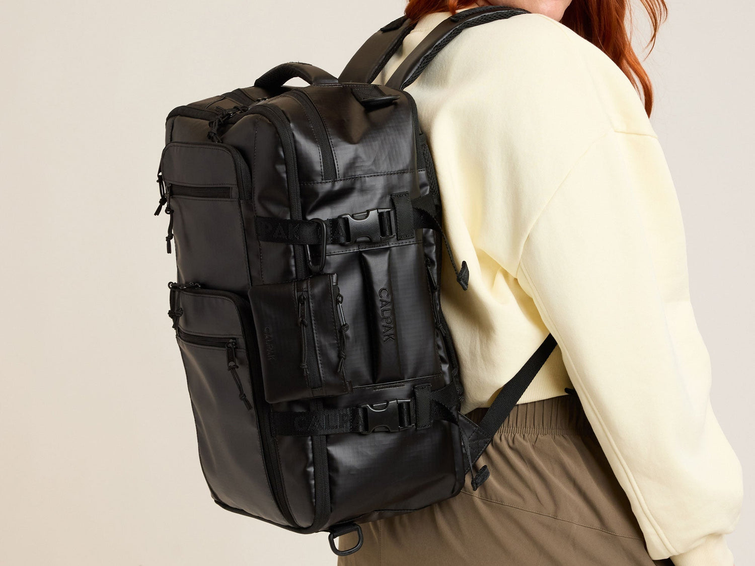 CALPAK Terra 26L Laptop Duffel Backpack in matte black Matte Obsidian worn by a model in a yellow sweater against a tan background