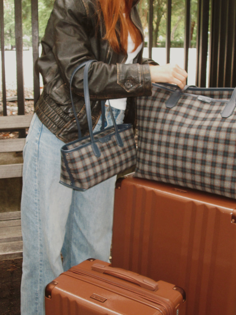 A person in a black leather jacket and light blue jeans carries a small CALPAK plaid tote bag next to larger matching plaid and brown Ambeur luggage bags.