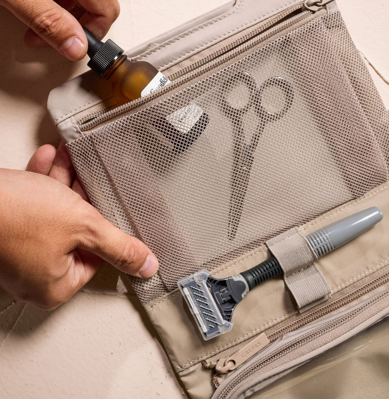 Person opening a beige CALPAK Fit Hanging Dopp Kit with grooming items including a razor and scissors.