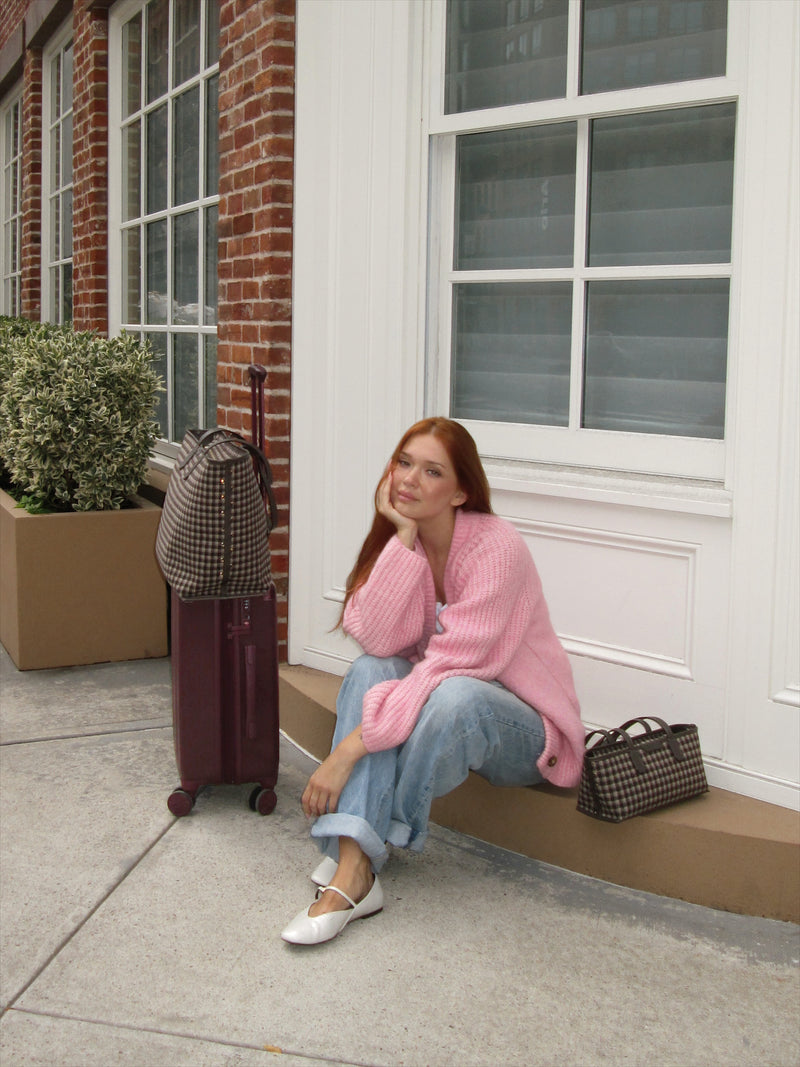 A woman with red hair sits on a concrete ledge outside a building, wearing a chunky pink knit sweater, light blue jeans, and white Mary Jane flats. A dark burgundy Ambeur Carry-On Luggage stands on the sidewalk next to her, with a plaid Colette Tote Bag resting on top and a small plaid handbag beside her.