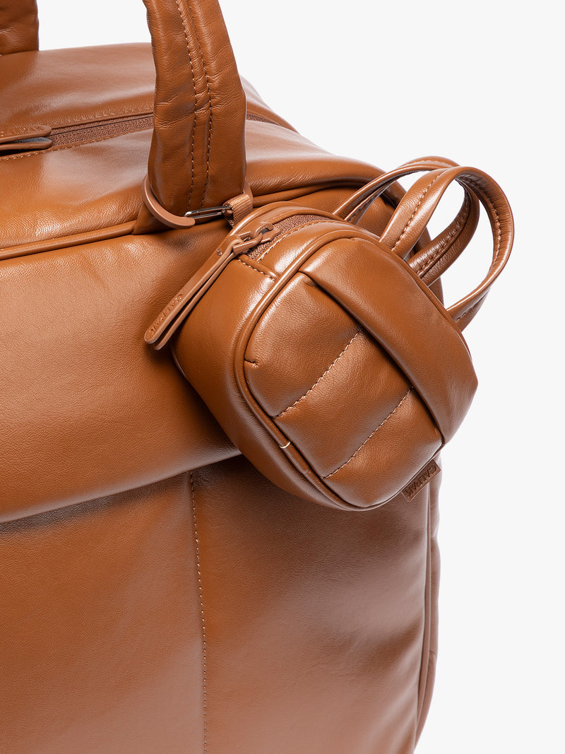 A close-up of a light Honey Brown vegan leather top-handle CALPAK Luka Zip Bag Charm with a zippered front pocket and a small side strap  with a matching small zippered pouch attached near the handle, shown against a clean white background
