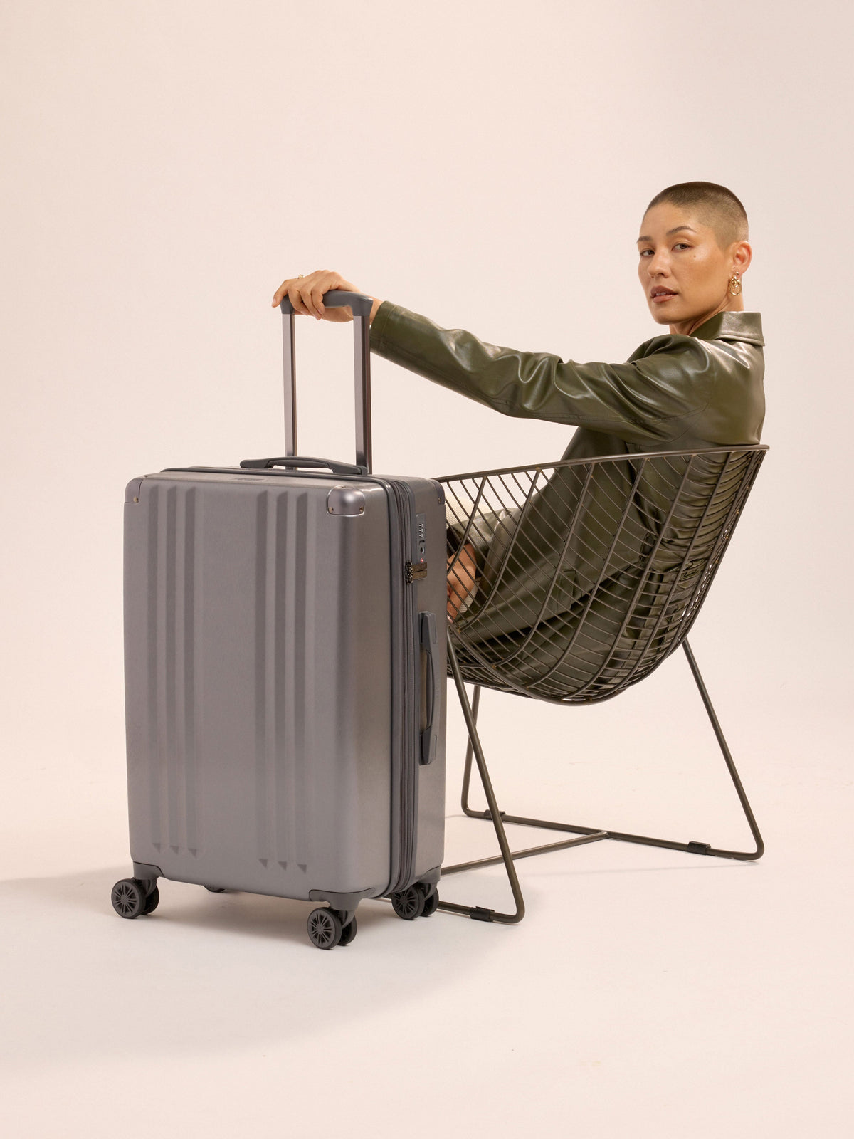 Model sitting besides CALPAK Ambeur Medium Luggage while holding telescopic trolley handle in metallic gunmetal gray; LAM1024-GUNMETAL