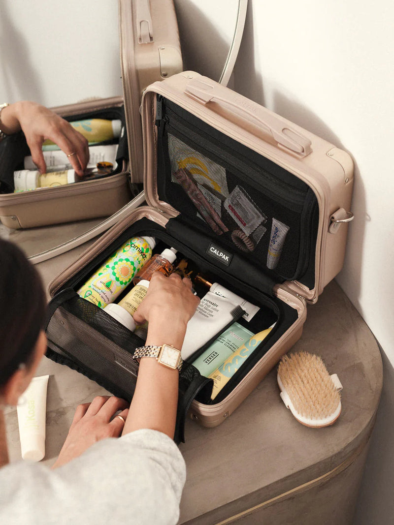 Open view of the CALPAK Ambeur Vanity Case in gold. Beauty products are being placed inside the mesh zippered compartments.