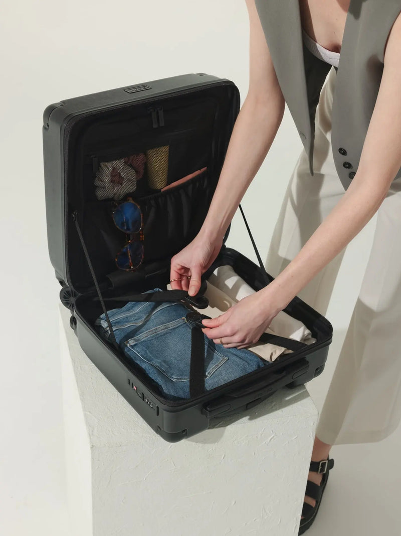 The CALPAK Ambuer Mini Carry-On suitcase in a stylish black finish used by a model packing. This compact rolling a mesh zippered compartment wirh compression straps for security.