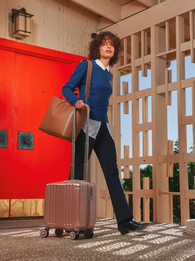 The CALPAK Ambuer Mini Carry-On suitcase in a stylish rose gold finish used by a model pulling it alongside her in an outdoor setting. This compact rolling suitcase has a glossy, smooth exterior with a metallic sheen. It includes a black retractable handle, four black swivel wheels for easy manoeuvrability.