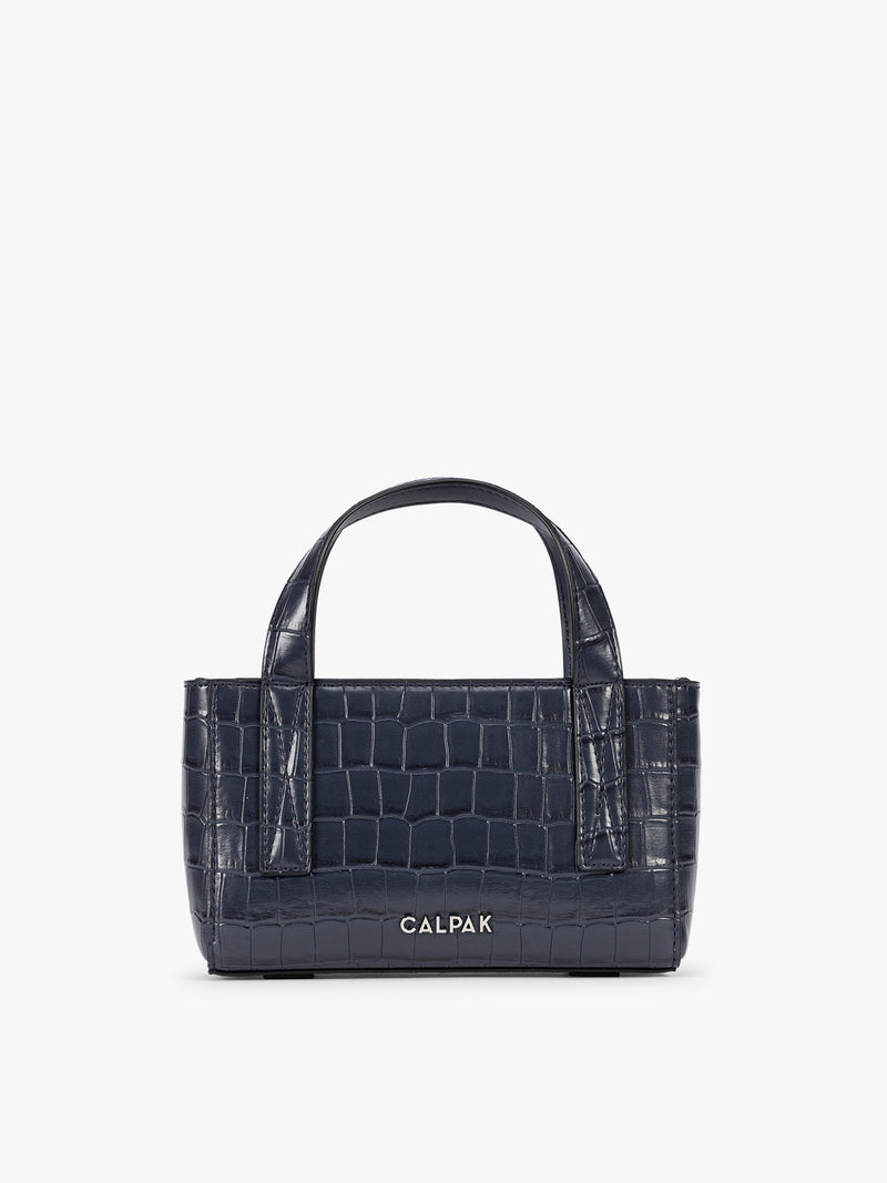 A CALPAK Haven Mini Crossbody Bag in Navy blue crocodile-embossed with top handles on a plain white background