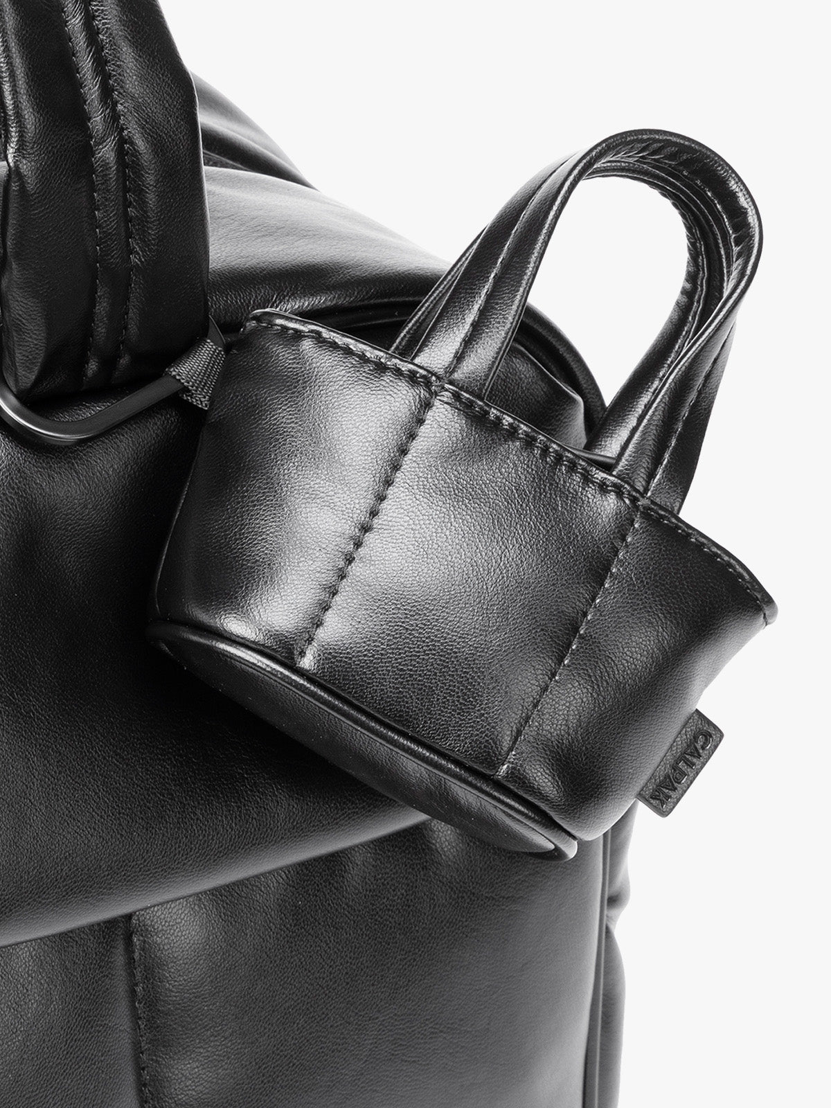 Closeup of a small Black CALPAK Luka Tote Bag Charm with dual top handles and a black carabiner clip attached to the side, shown against a plain white background; AMT2601-BLACK-PU