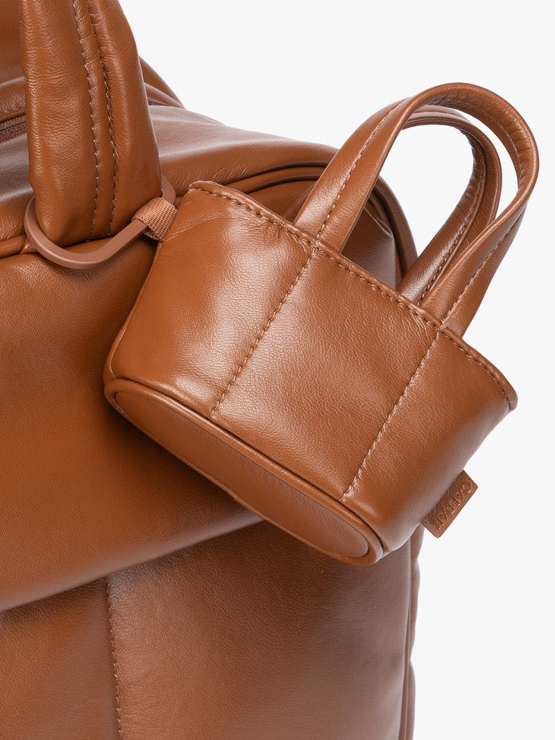 Closeup of a small light brown Honey Brown CALPAK Luka Tote Bag Charm with dual top handles and a black carabiner clip attached to the side, shown against a plain white background