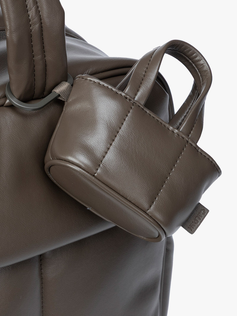 Closeup of A small dark brown Mushroom CALPAK Luka Tote Bag Charm with dual top handles and a black carabiner clip attached to the side, shown against a plain white background
