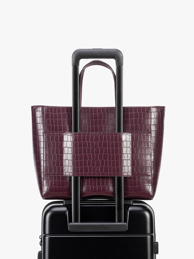 Backside luggage trolley sleeve of CALPAK Haven 15" Laptop Tote in deep red Dark Cherry crocodile-embossed with dual top handles and a matching shoulder strap against a plain white background