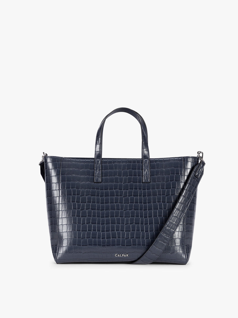 A navy CALPAK Haven 15" Laptop Tote in Navy blue crocodile-embossed with dual top handles and a matching shoulder strap against a plain white background