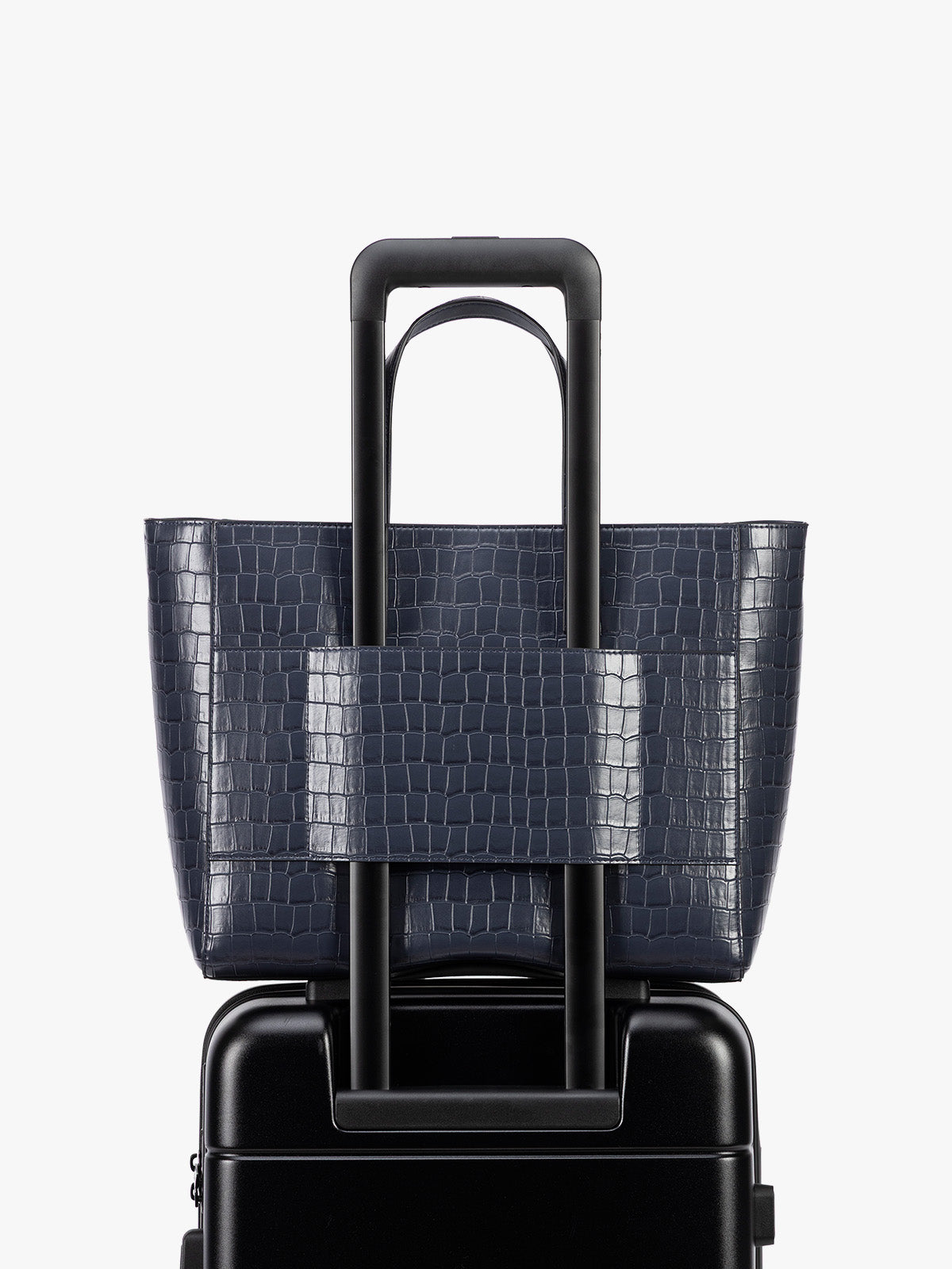 Backside luggage trolley sleeve of A navy CALPAK Haven 15" Laptop Tote in Navy blue crocodile-embossed with dual top handles and a matching shoulder strap against a plain white background; ATS2501-NAVY-FAUX-CROC