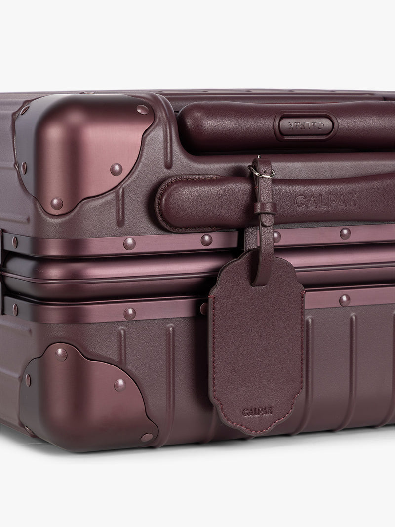 A close-up of a deep red hard-shell suitcase with protective corner bumpers, a deep red CALPAK Vault luggage tag with a scalloped border and a matching black strap, shown against a plain white background