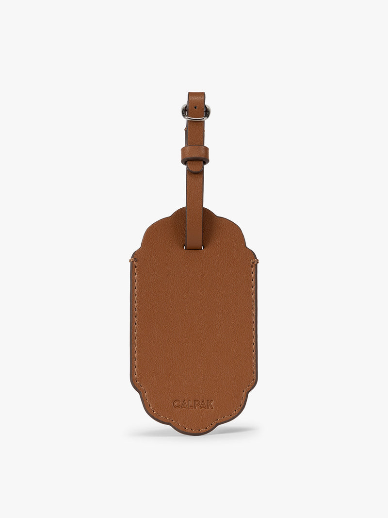 Backside of a honey brown CALPAK Vault luggage tag with a scalloped border and a matching honey brown strap, shown against a plain white background