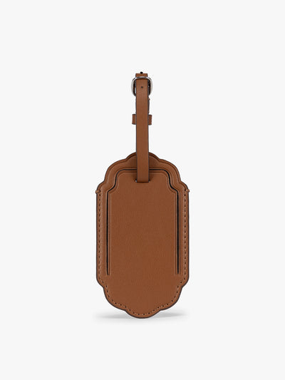 A honey brown CALPAK Vault luggage tag with a scalloped border and a matching honey brown strap, shown against a plain white background; ATV2601-HONEY-BROWN view 1