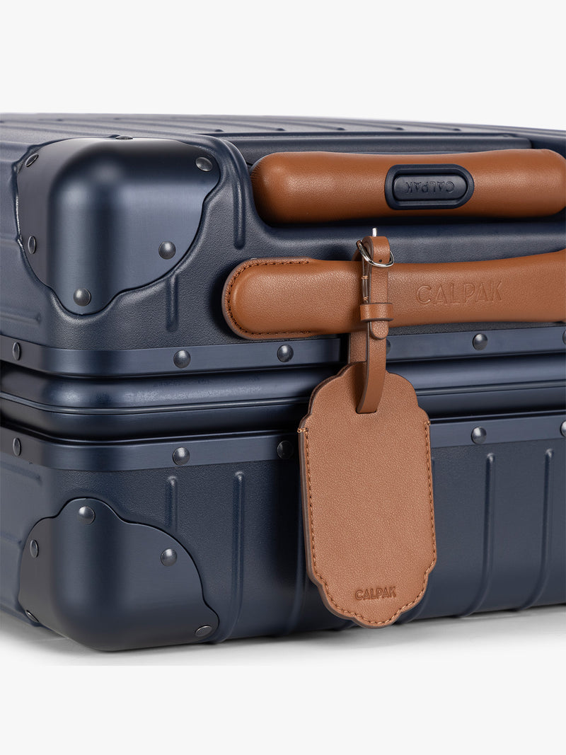 A close-up of a blue hard-shell suitcase with protective corner bumpers, a honey brown CALPAK Vault luggage tag with a scalloped border and a matching black strap, shown against a plain white background