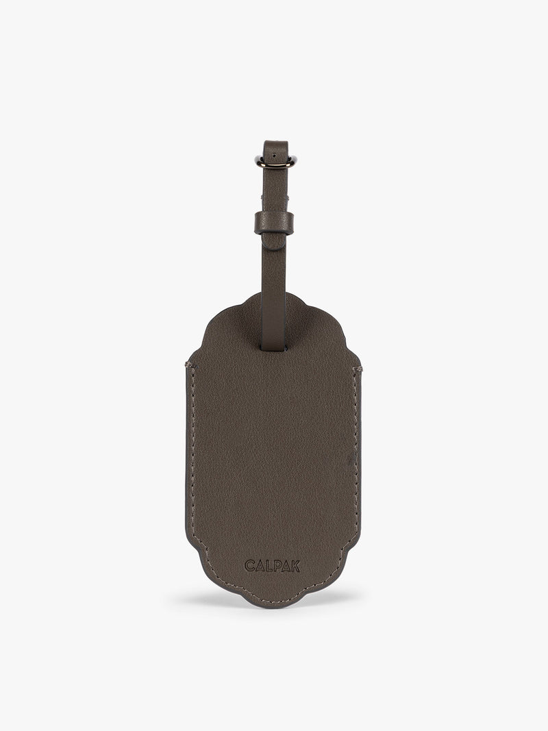 Backside of a dark brown CALPAK Vault luggage tag with a scalloped border and a matching dark brown strap, shown against a plain white background