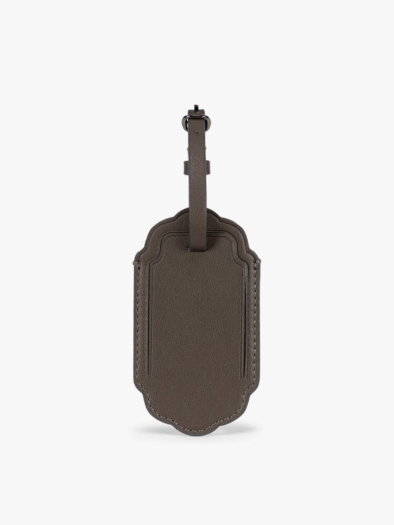 A dark brown CALPAK Vault luggage tag with a scalloped border and a matching dark brown strap, shown against a plain white background