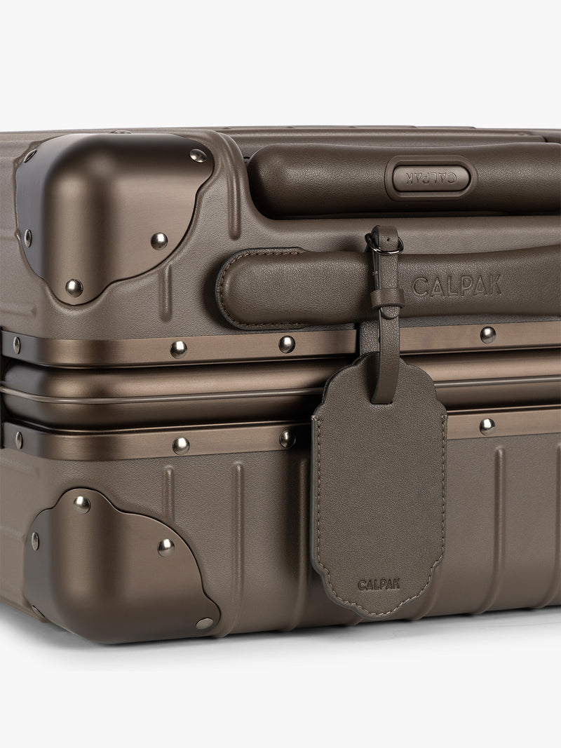 A close-up of a brown hard-shell suitcase with protective corner bumpers, a brown CALPAK Vault luggage tag with a scalloped border and a matching black strap, shown against a plain white background