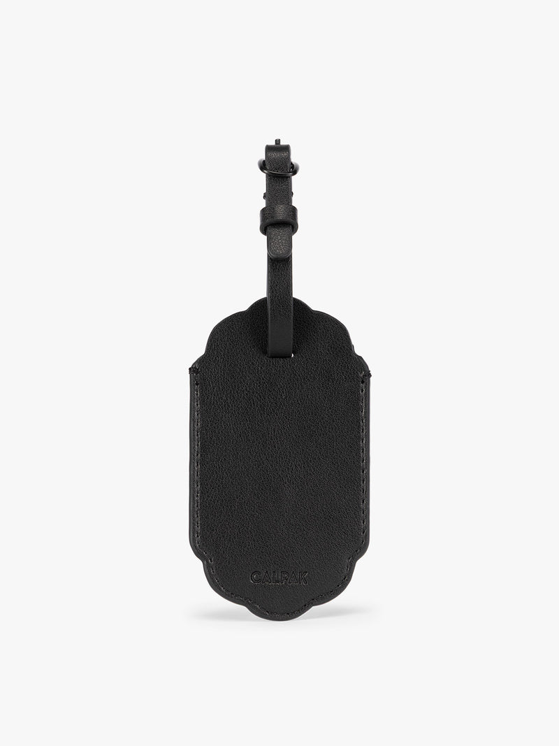 Backside of a black CALPAK Vault luggage tag with a scalloped border and a matching black strap, shown against a plain white background