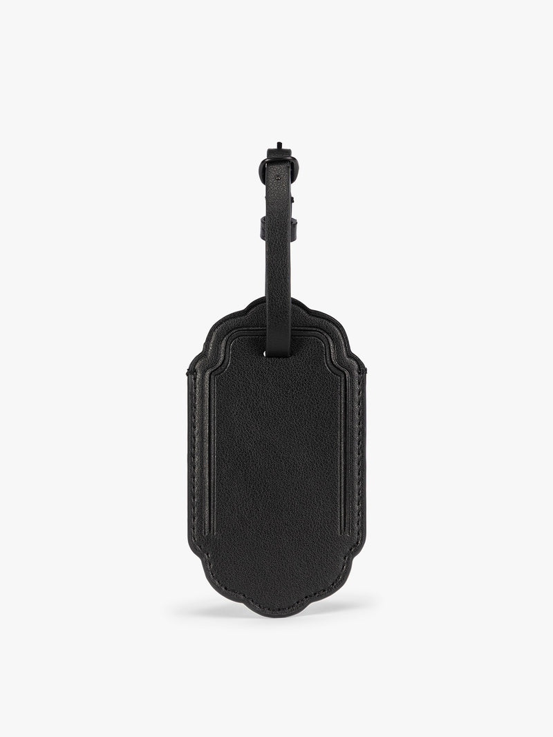 A black CALPAK Vault luggage tag with a scalloped border and a matching black strap, shown against a plain white background