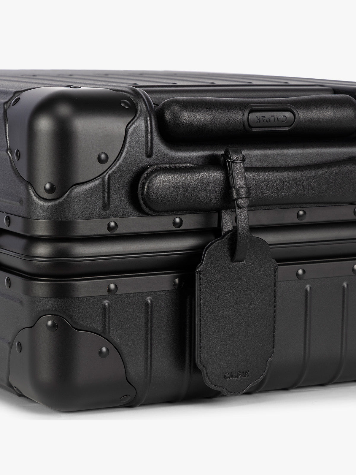 A close-up of a black hard-shell suitcase with protective corner bumpers, a black CALPAK Vault luggage tag with a scalloped border and a matching black strap, shown against a plain white background; ATV2601-BLACK