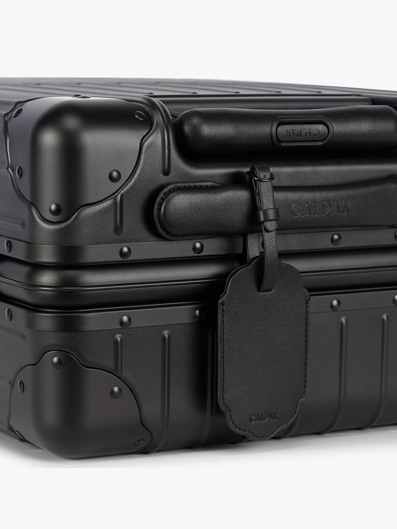 A close-up of a black hard-shell suitcase with protective corner bumpers, a black CALPAK Vault luggage tag with a scalloped border and a matching black strap, shown against a plain white background