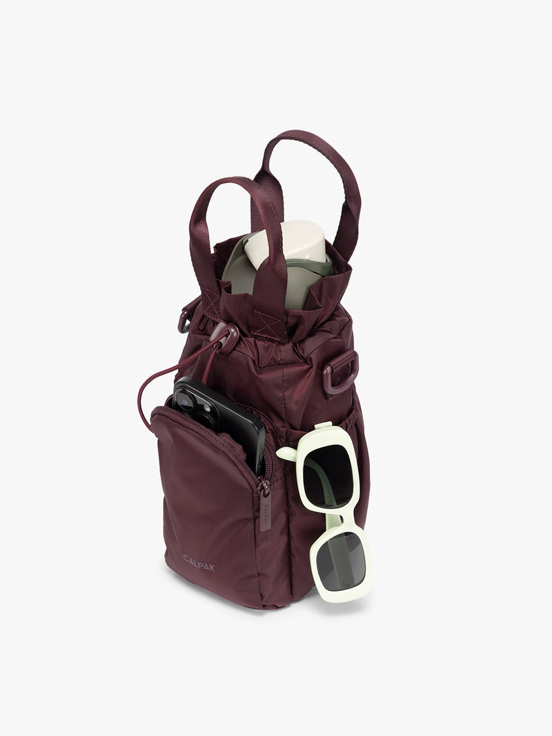 Water bottle, phone, and sunglasses in the Burgundy Dark Cherry CALPAK Water Bottle Holder with top handles, drawstring top, front zippered pocket, and a detachable shoulder strap that is expandable