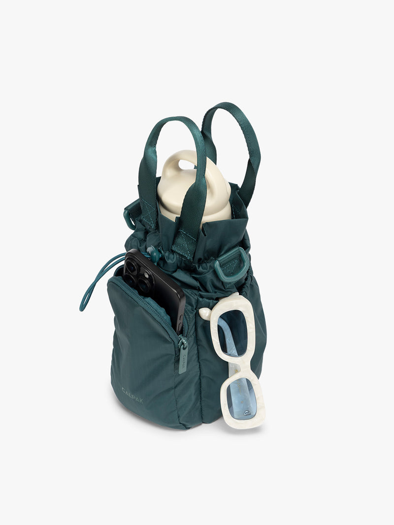 Water bottle, phone, and sunglasses in the dark green teal Kale CALPAK Water Bottle Holder with top handles, drawstring top, front zippered pocket, and a detachable shoulder strap with expandable top