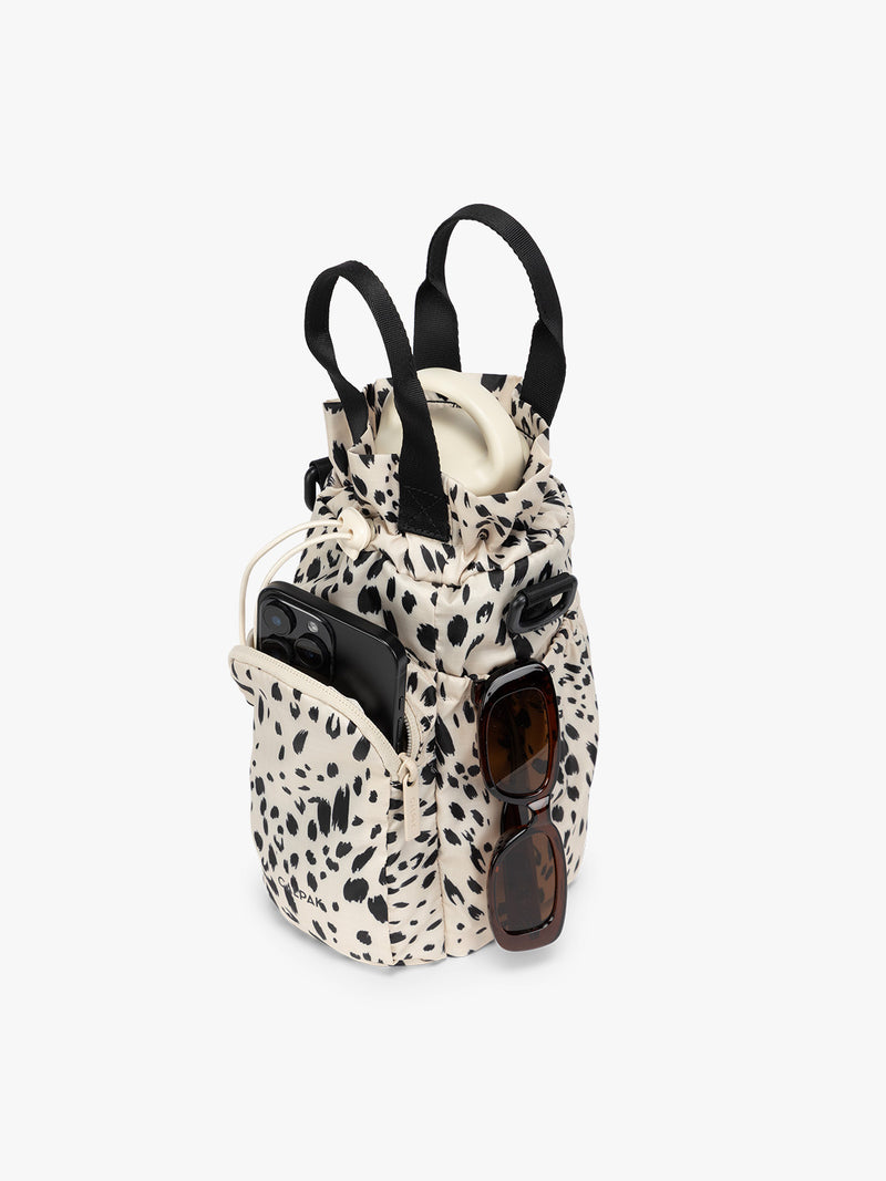 Water bottle, phone, and sunglasses in the white and black animal print Snow Leopard CALPAK Water Bottle Holder with top handles, drawstring top, front zippered pocket, and a detachable shoulder strap that is expandable
