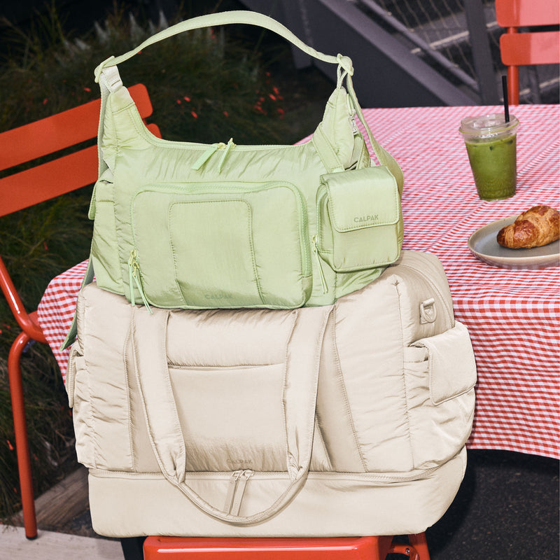 CALPAK Crossbody Diaper Bag in light green Lime on top of the CALPAK Weekender Duffel in white Oatmeal on a red stool with a table in the background