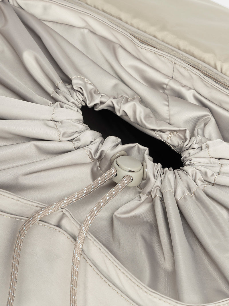 Close-up of a gray drawstring bag with a button closure of CALPAK Fit Gym Backpack in beige Greige with white background