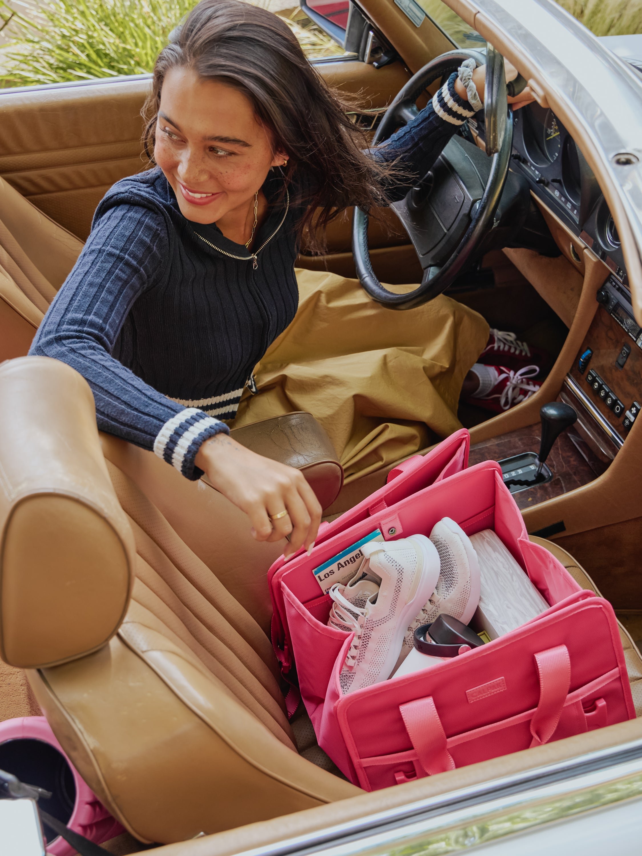 Model in convertible car with collapsed CALPAK Car Organizer in pink dragonfruit in the passenger seat; ACO2001-DRAGONFRUIT
