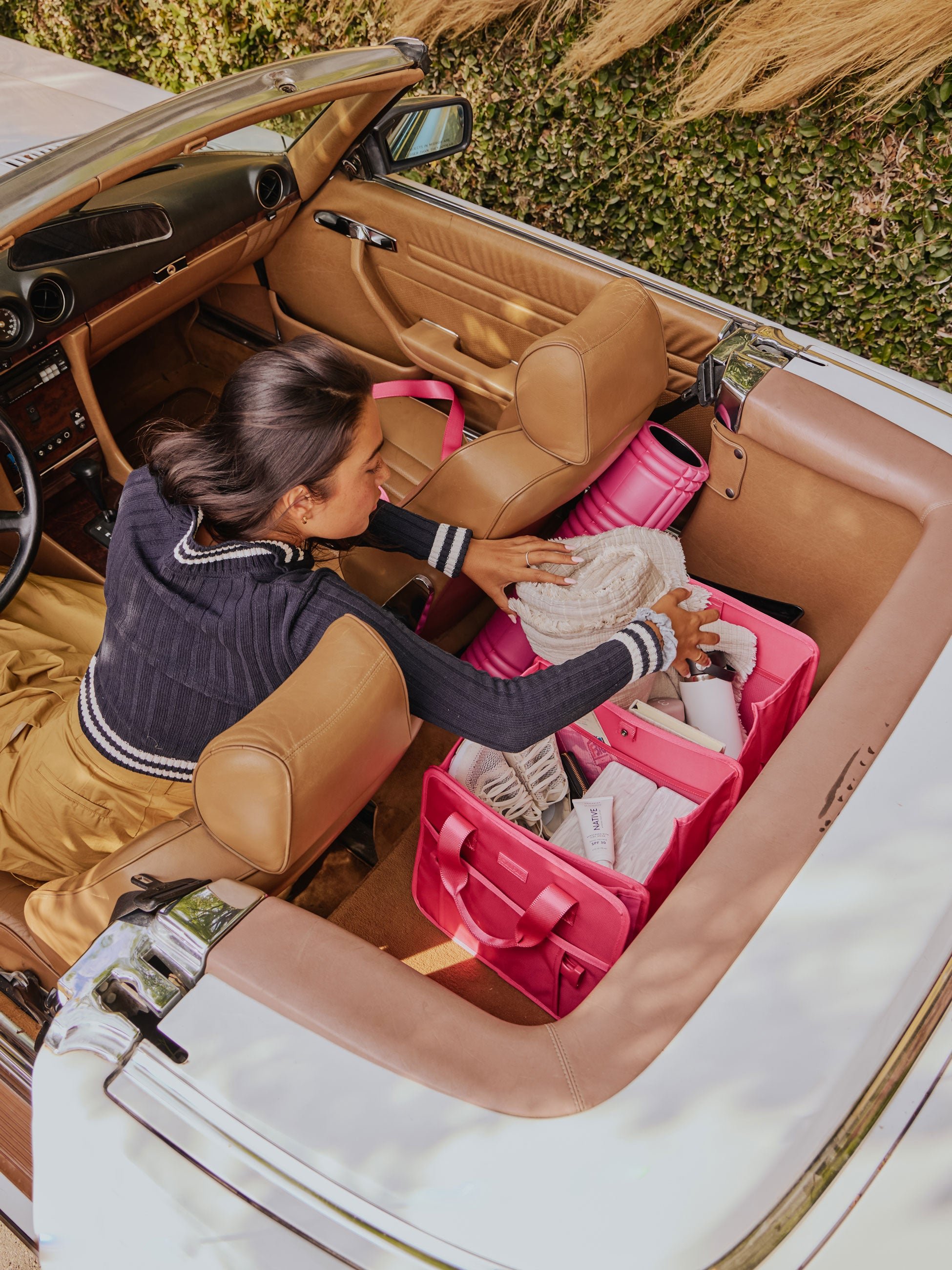 Model sitting in convertible car reaching towards the backseat for blanket in her CALPAK Car Organizer in dragonfruit