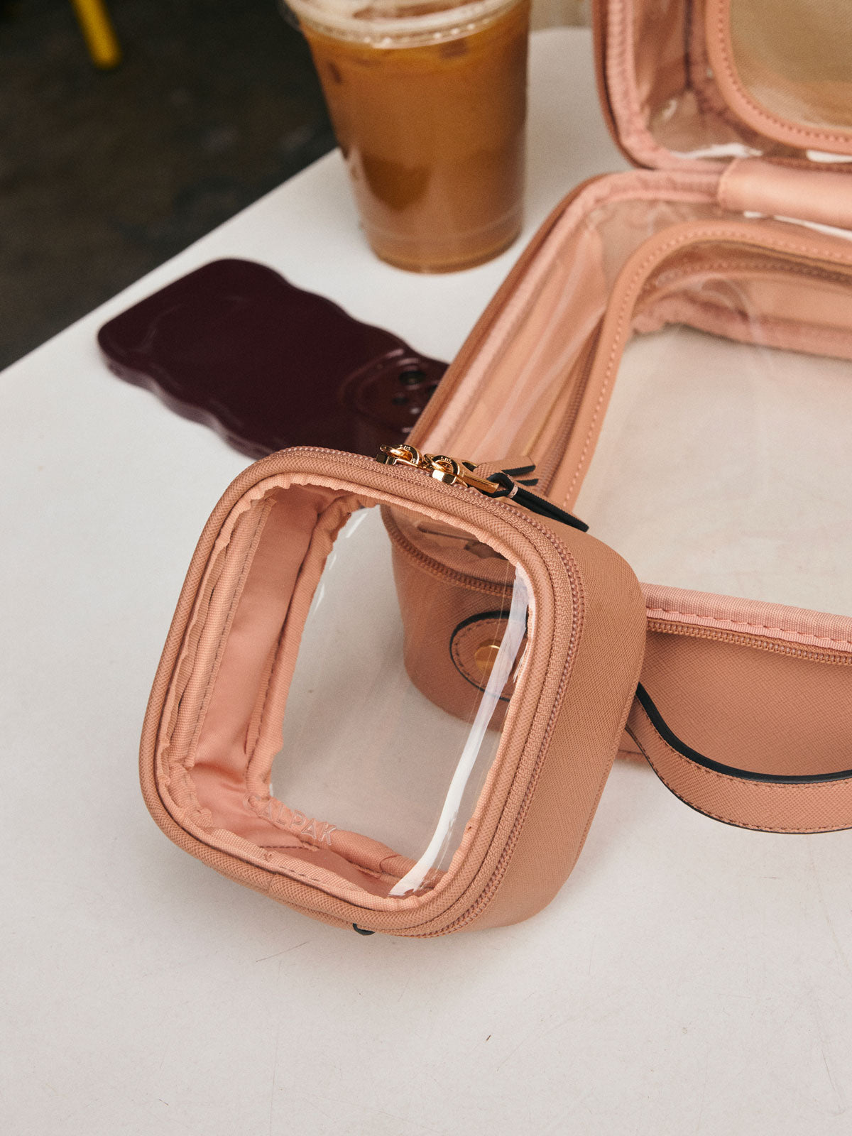 CALPAK Tiny Clear Cosmetic Case in light brown Caramel on a white table with other CALPAK clear cosmetic case, coffee, and a phone  in the background; CCT2501-CARAMEL