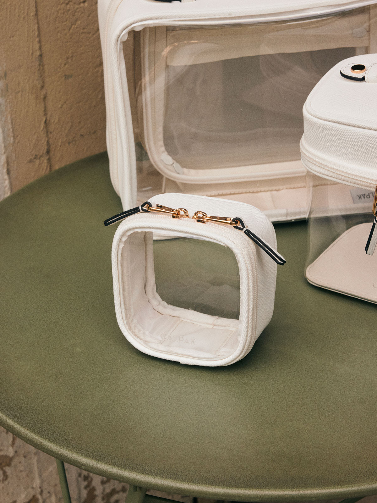 CALPAK Tiny Clear Cosmetic Case in White on a green table with other CALPAK clear cosmetic cases in the background; CCT2501-WHITE