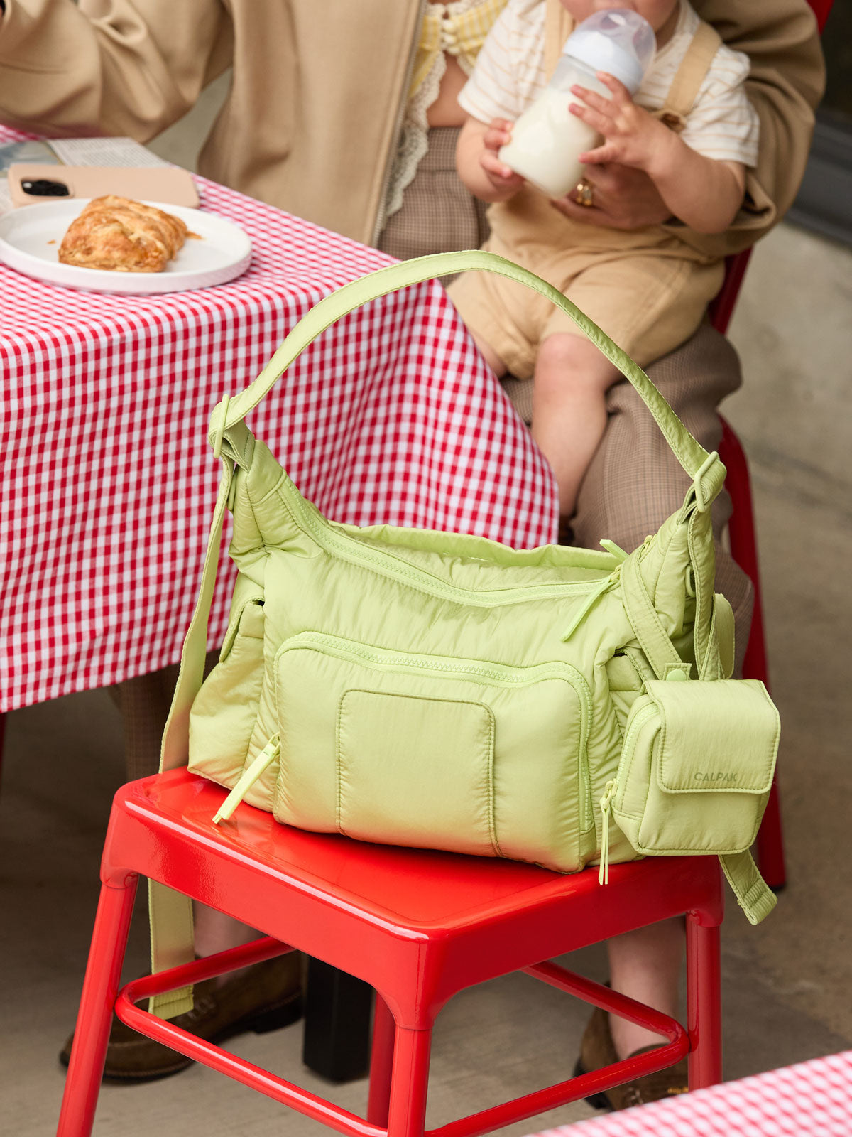 CALPAK Crossbody Diaper Bag in brown light green Lime on a red stool with a mom and baby in the background; CDB2501-LIME
