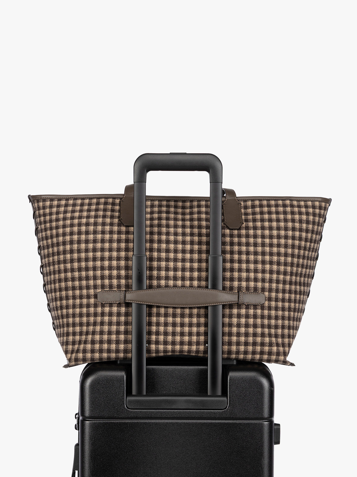 A brown and beige checkered tote bag with dark brown leather handles, resting on top of a black rolling suitcase against a plain white background; CLCT2601-MUSHROOM-PLAID