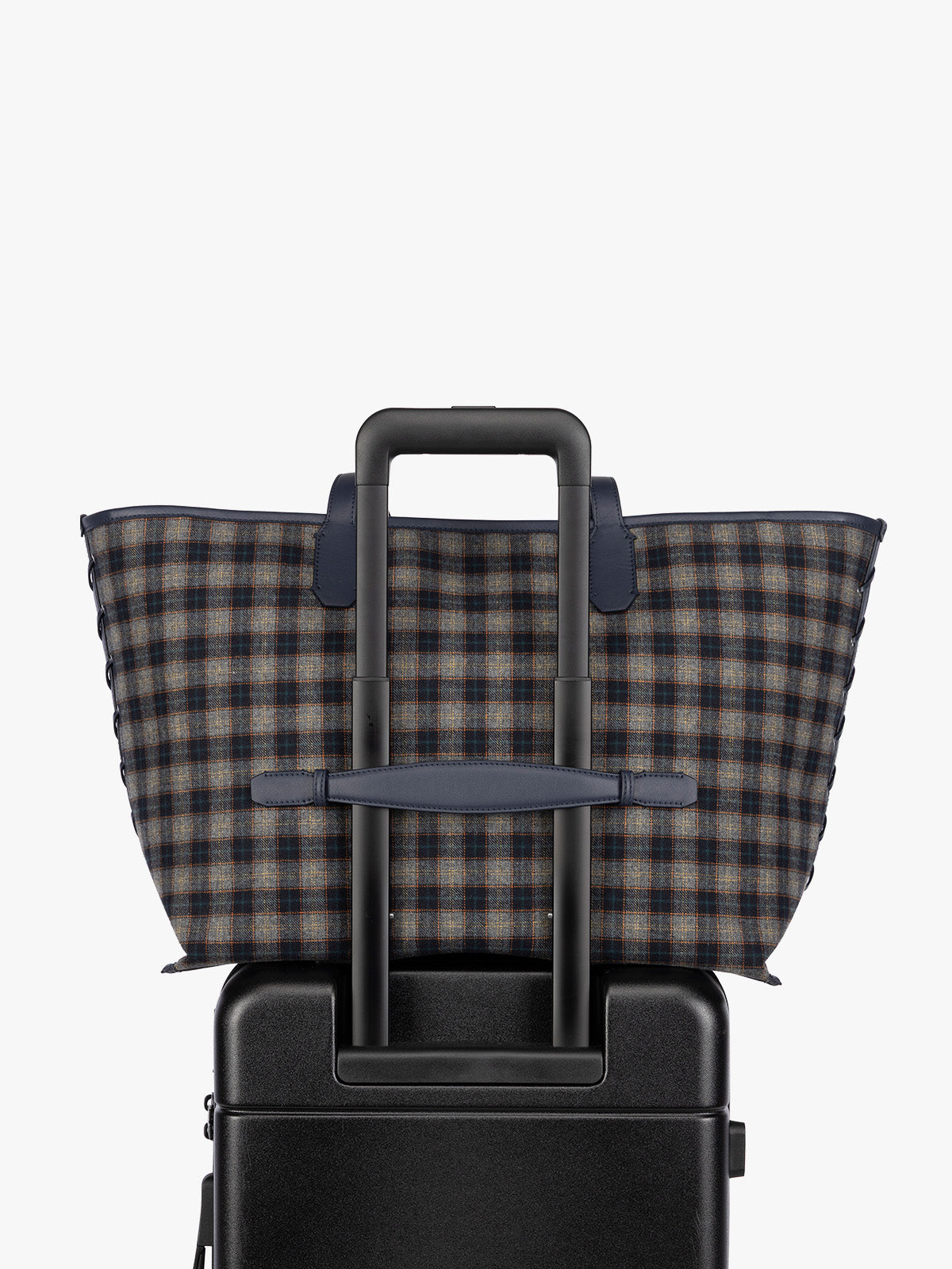 A CALPAK Colette 15" Laptop tote bag with a brown and navy blue plaid pattern and dark navy blue handles, shown resting on top of a black rolling suitcase against a plain light gray background ; CLCT2601-NAVY-PLAID, 